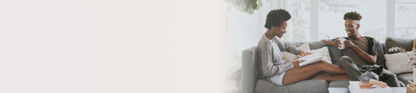 A Black woman reading a book and a Black man drinking from a white mug sitting on a grey couch